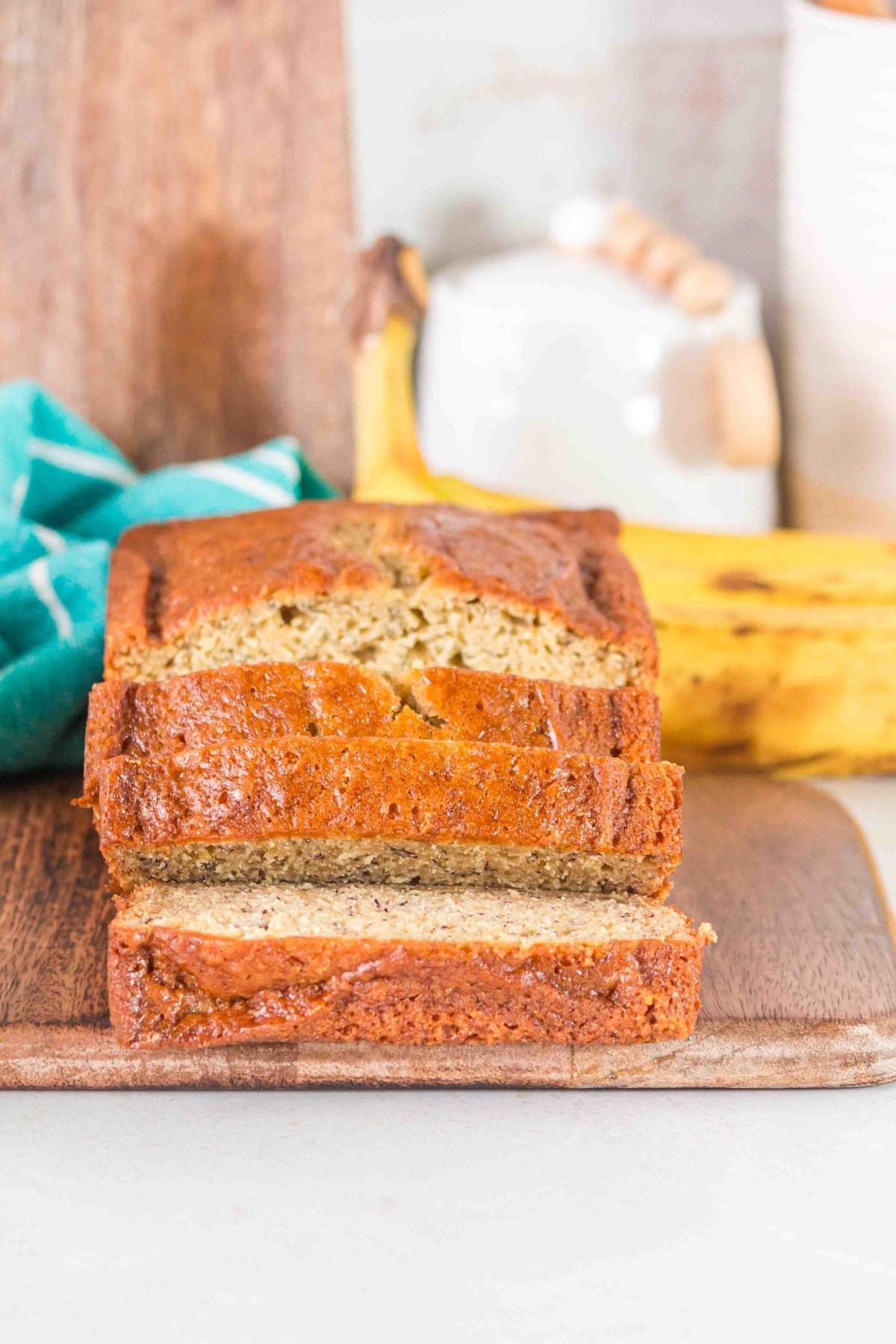 A loaf of brown butter banana bread, sliced perfectly on a wooden board, sits invitingly with a ripe banana nearby and a blue cloth elegantly draped in the background.