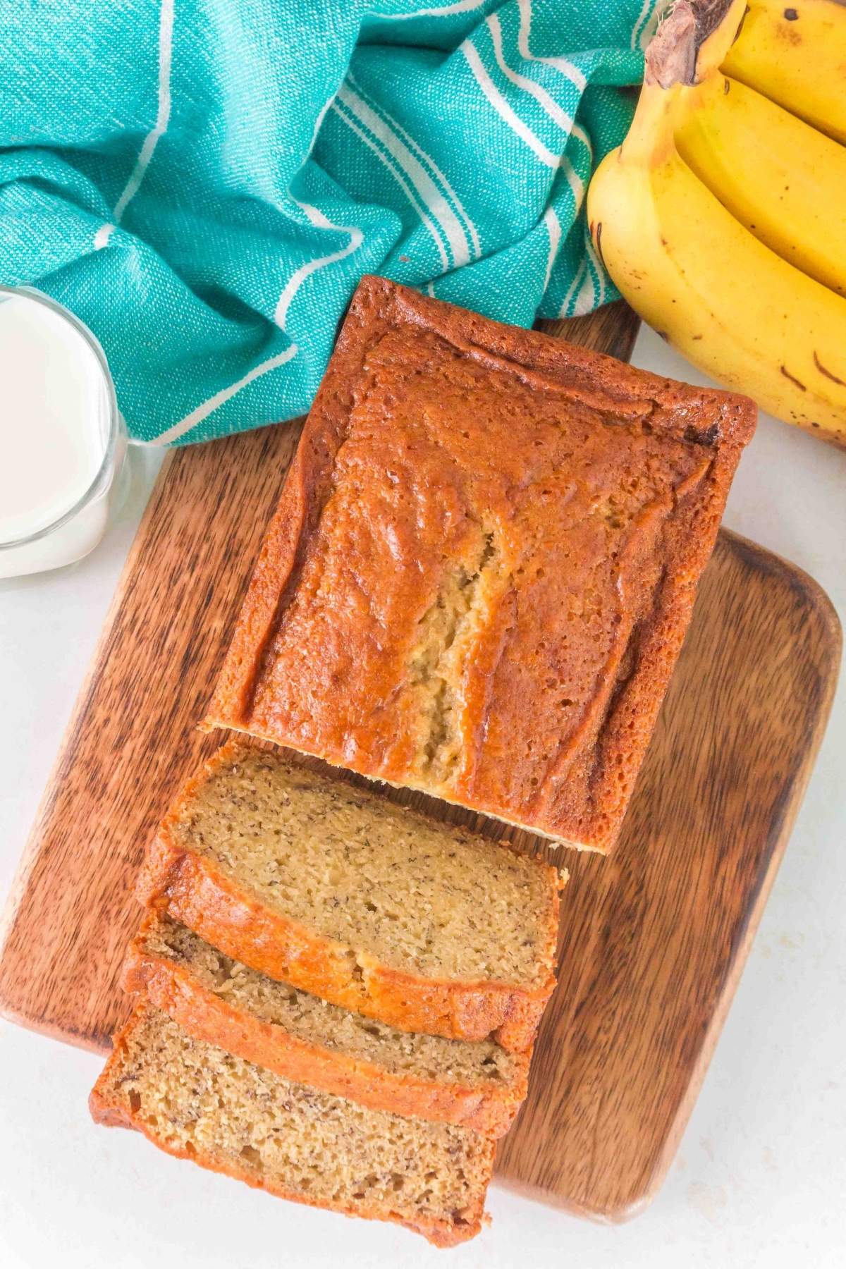 A loaf of brown butter banana bread with slices on a wooden board sits next to a bunch of bananas, a glass of milk, and a green striped cloth.