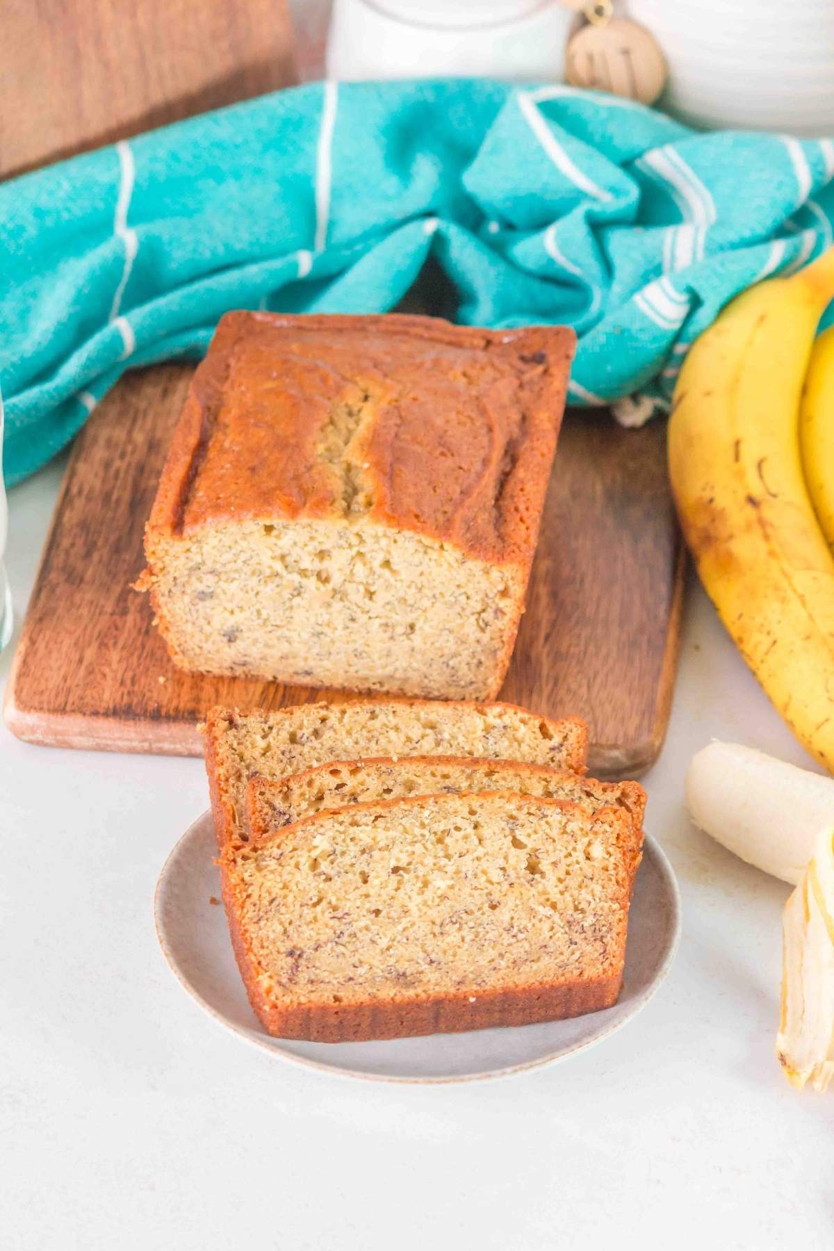 Sliced brown butter banana bread on a plate with bananas and a teal cloth in the background.