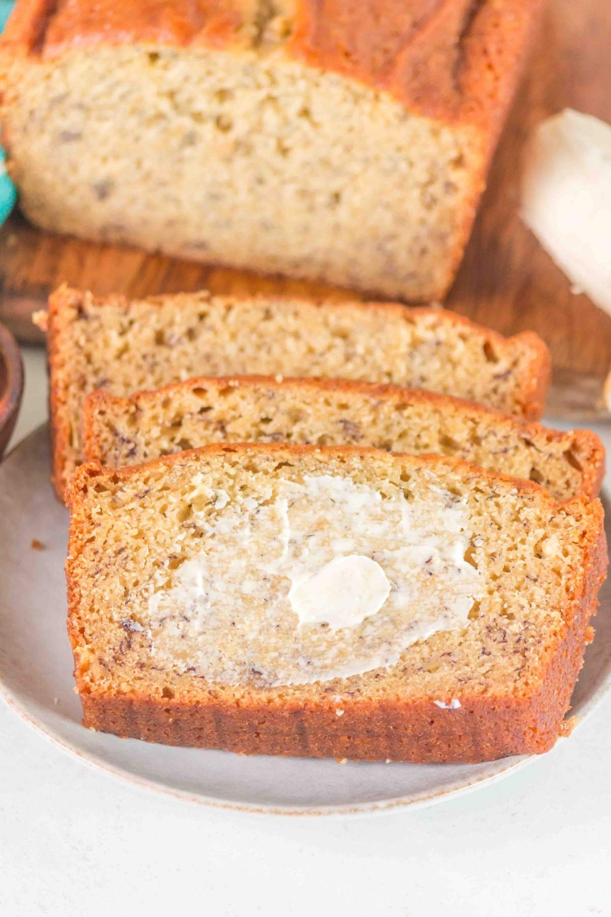 Slices of brown butter banana bread rest on a plate, with one slice crowned with a pat of butter. In the background, a partially visible loaf adds to the cozy scene.