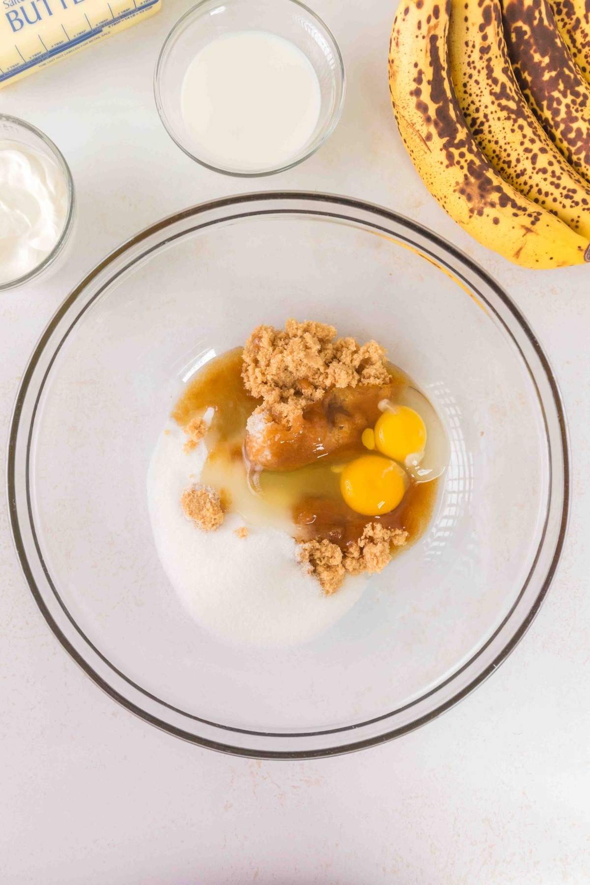 A glass bowl contains brown sugar, white sugar, two eggs, and vanilla extract. Nearby are ripe bananas, sour cream, milk, and the key to rich flavor—brown butter—awaiting their transformation into delicious banana bread.
