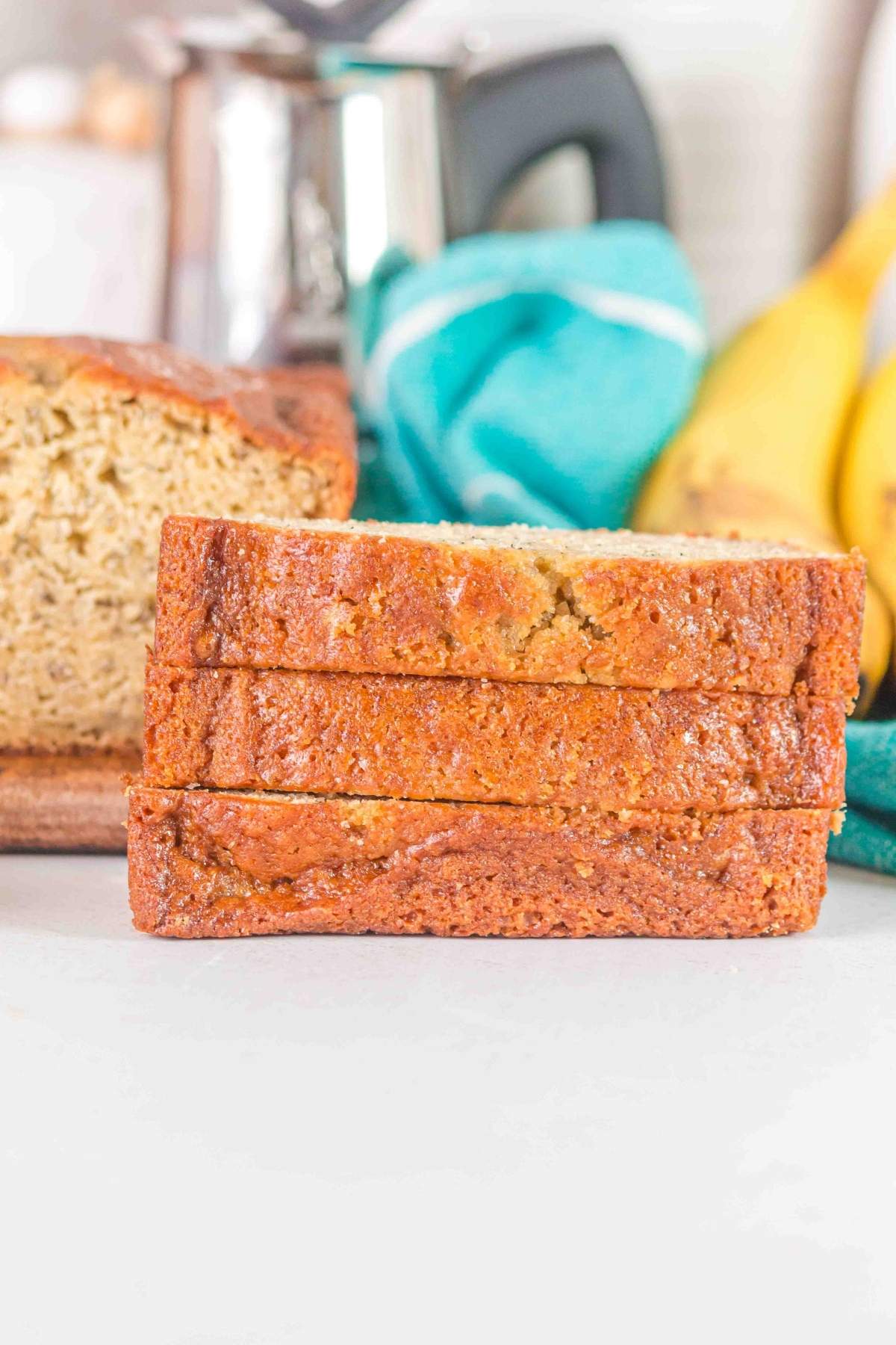 Three slices of brown butter banana bread stacked on a white surface with a loaf, bananas, a towel, and a metal coffee pot in the background.