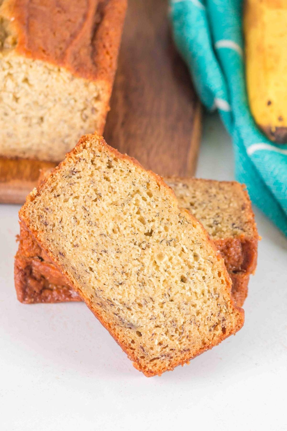 Close-up of two slices of brown butter banana bread on a wooden board, with a blue cloth and a ripe banana in the background.