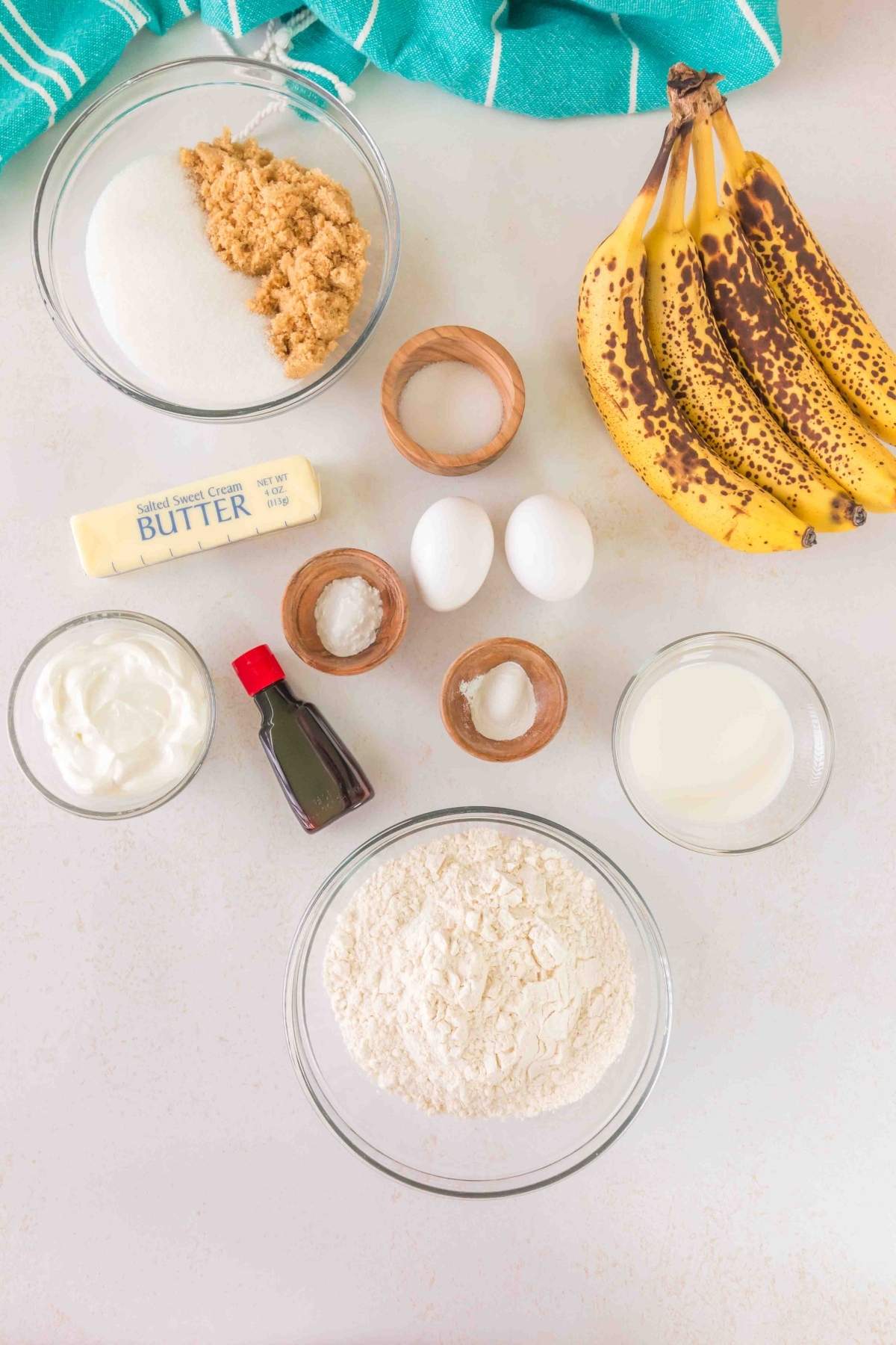Ingredients for brown butter banana bread on a countertop: ripe bananas, nutty brown butter, sugars, eggs, milk, flour, baking powder, salt, vanilla extract, and yogurt.