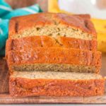 Brown butter banana bread, perfectly sliced on a wooden board, with a teal-striped towel and fresh bananas artfully arranged in the background.