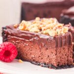 A luscious slice of chocolate cheesecake topped with chopped nuts sits elegantly on a white plate, surrounded by vibrant, fresh raspberries.