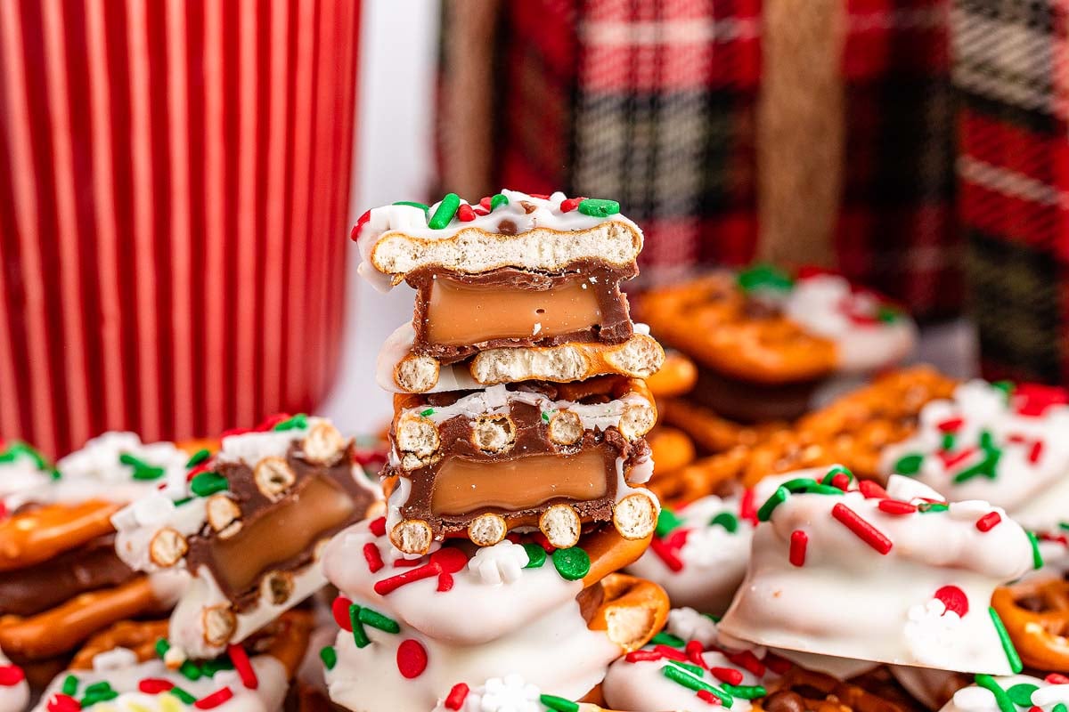Close-up of stacked Christmas Rolo pretzel sandwiches with caramel and chocolate, coated in white icing and festive sprinkles. Plaid and striped fabrics provide a cozy backdrop.