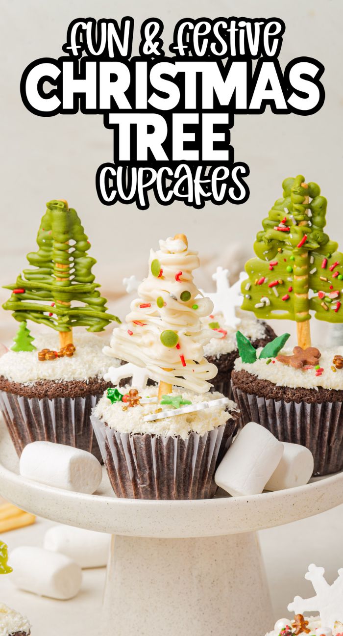 Christmas tree cupcakes topped with festive decorations and sprinkles are displayed on a white cake stand, surrounded by fluffy marshmallows.