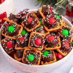 A bowl of holiday-themed treats features Christmas Rolo pretzel bites topped with chocolate, red and green candies, and colorful sprinkles. Additional ingredients and decorations are in the background, creating a festive atmosphere.