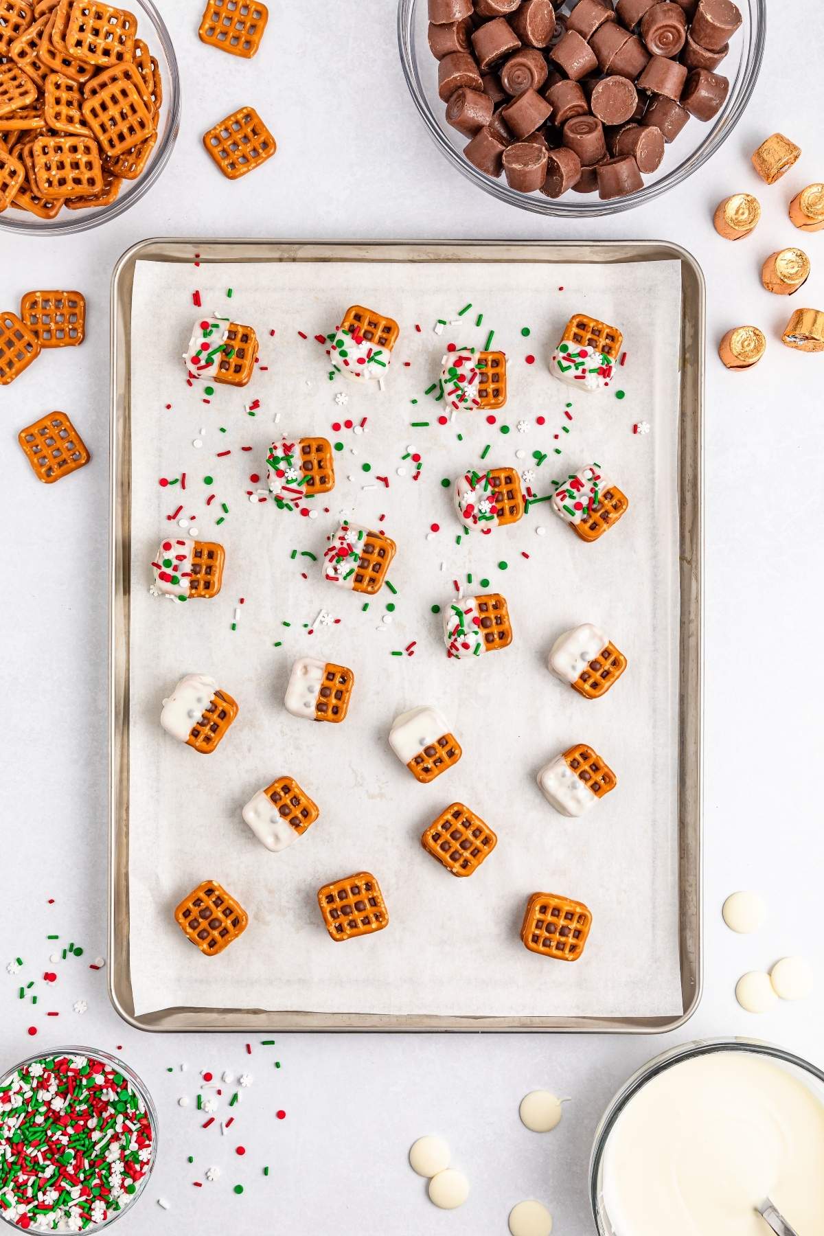 A baking sheet with pretzels, some dipped in white chocolate and sprinkles. Nearby, bowls brimming with Rolos and candy accompany a glass of milk, ready to delight any sweet tooth.