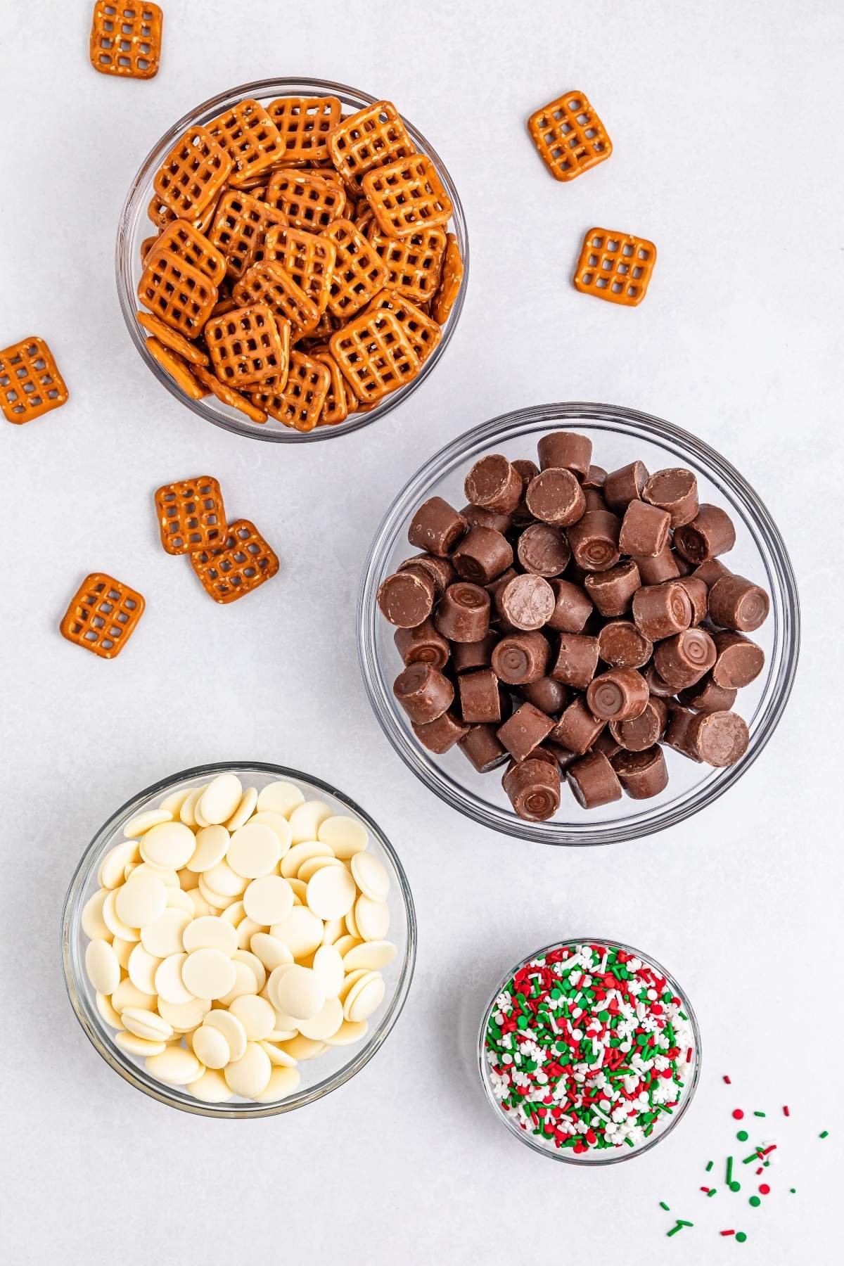 Four bowls with ingredients for Christmas Rolo pretzel sandwiches: pretzels, chocolate candies, white chocolate wafers, and festive red, green, and white sprinkles. Loose pretzels scattered around on a white surface.