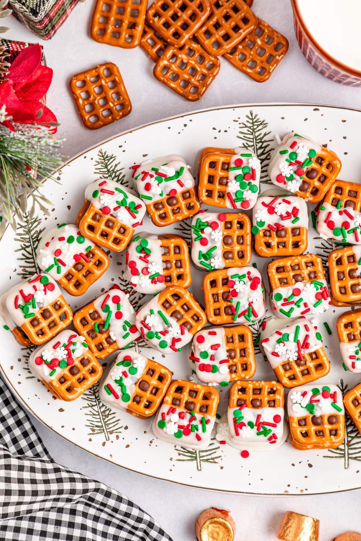 A plate of Christmas Rolo pretzel sandwiches, adorned with white icing and sprinkles, sits on a festive holiday-themed dish. Nearby are more pretzels and a piece of checkered fabric.