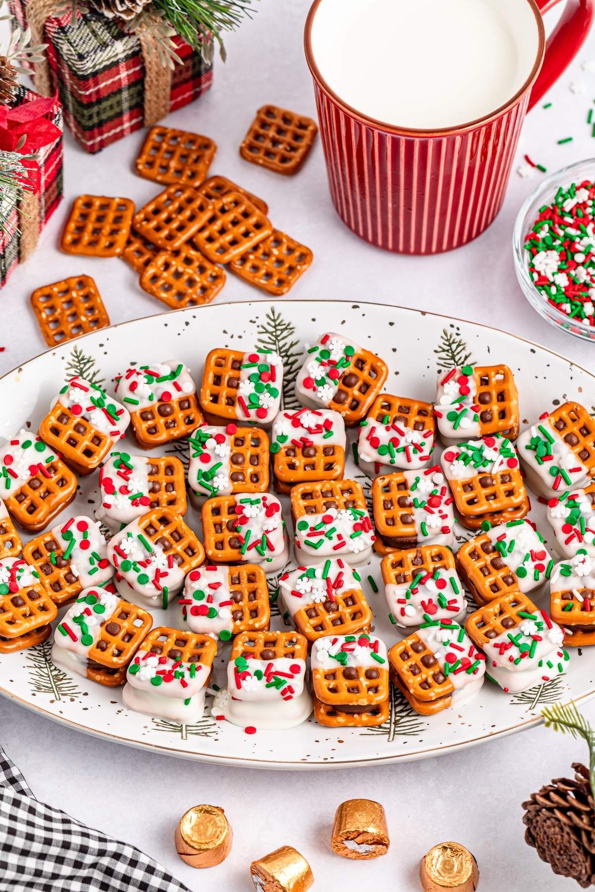 A plate of Christmas Rolo pretzel sandwiches topped with white chocolate and red, green sprinkles sits invitingly. Nearby, a mug of milk, scattered pretzels, and festive holiday decor complete the cheerful scene.