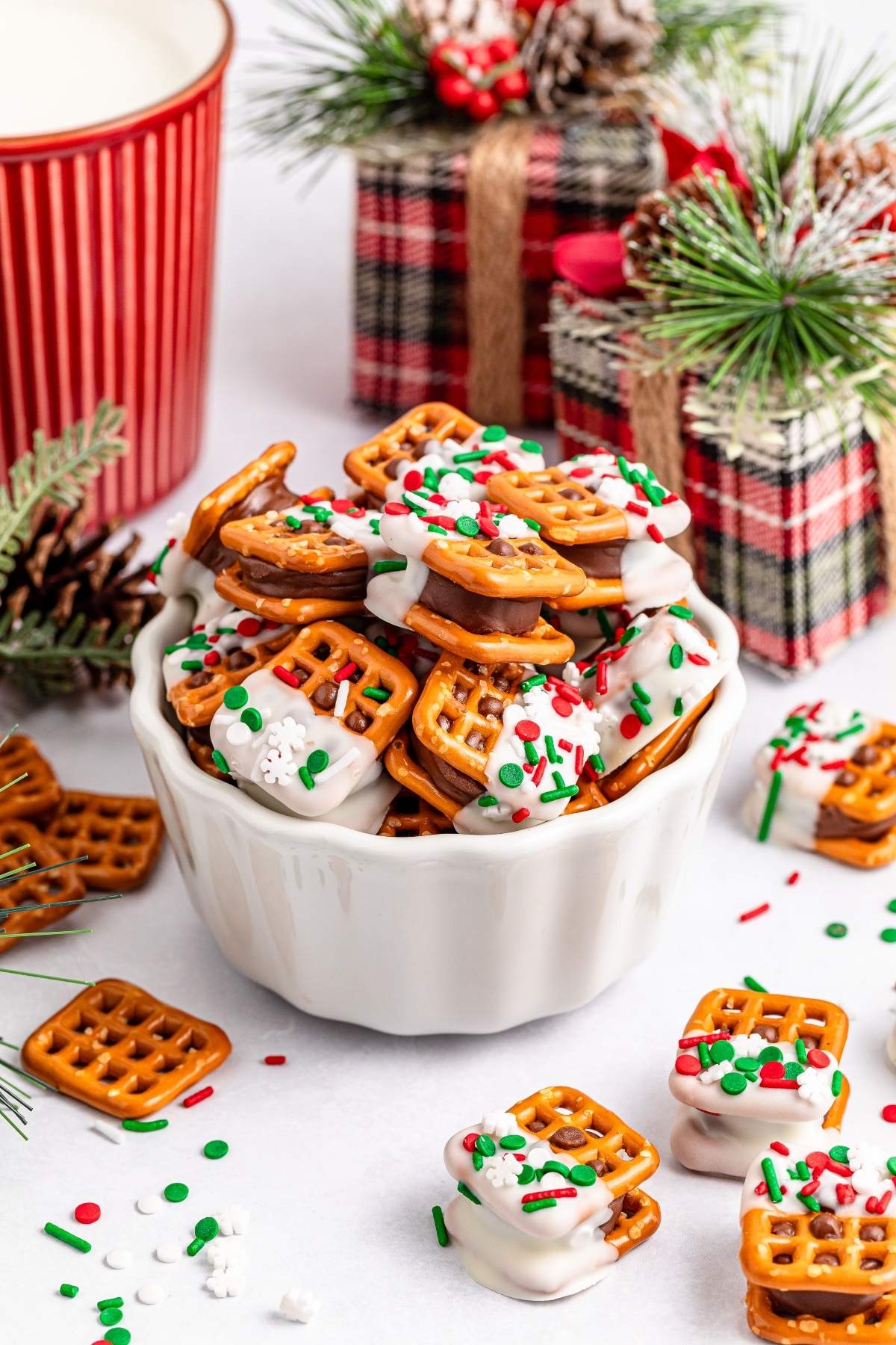 A bowl of white chocolate-coated Rolo pretzel sandwiches, adorned with red and green sprinkles, sits surrounded by festive decorations and gift boxes.