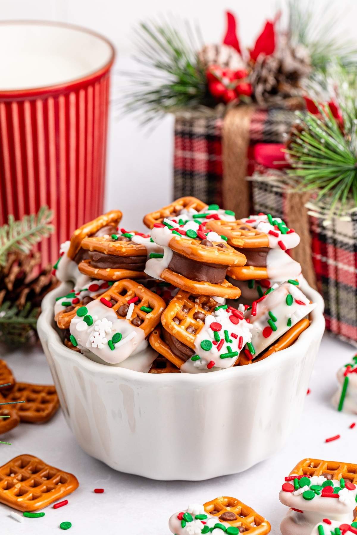 A white bowl brimming with Christmas Rolo pretzel sandwiches coated in white chocolate and adorned with red and green sprinkles. Festive decorations and a red striped mug accentuate the holiday spirit in the background.