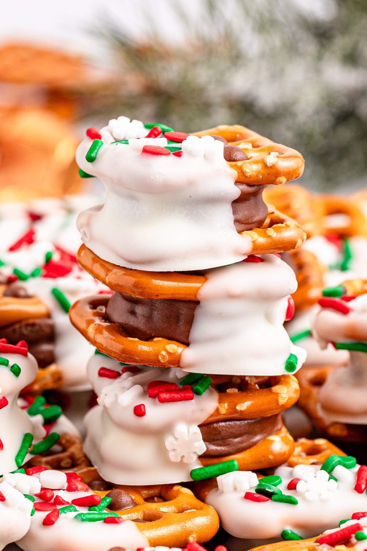 A festive stack of pretzel sandwiches dipped in white chocolate, adorned with red, green, and white sprinkles. More pretzels can be seen enticingly in the background.