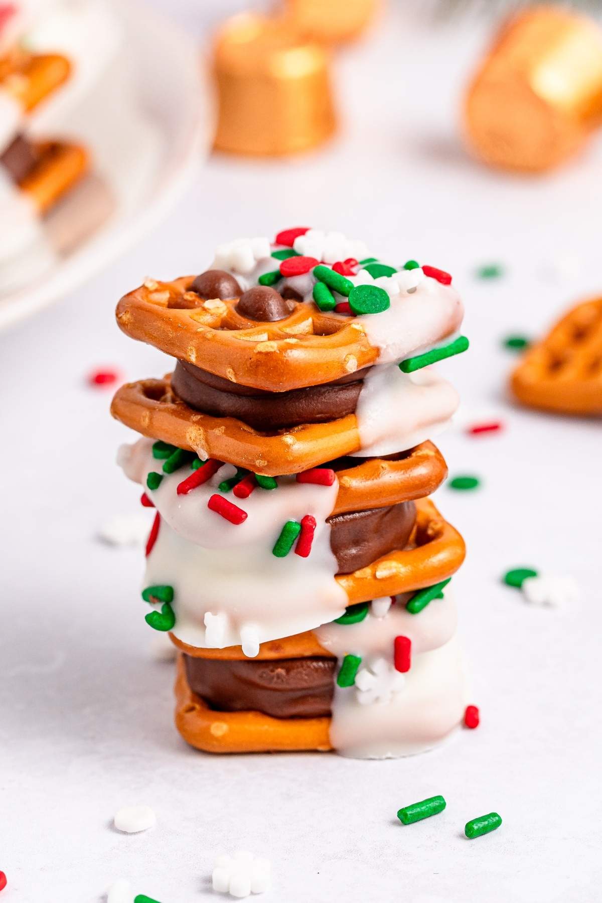 A festive stack of chocolate and white-coated pretzels sandwiches is topped with red, green, and white sprinkles on a pristine surface adorned with scattered sprinkles.