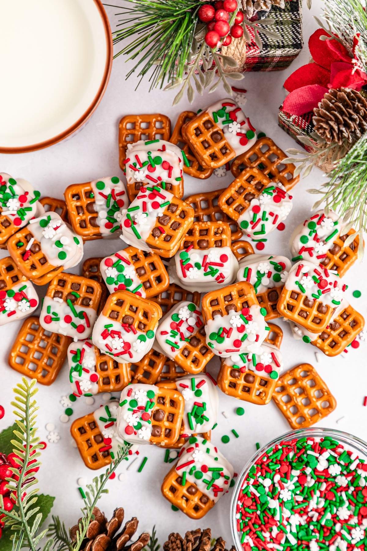Festive Christmas Rolo pretzel sandwiches topped with white icing and red, green sprinkles are artfully arranged on a table, surrounded by charming Christmas decorations.