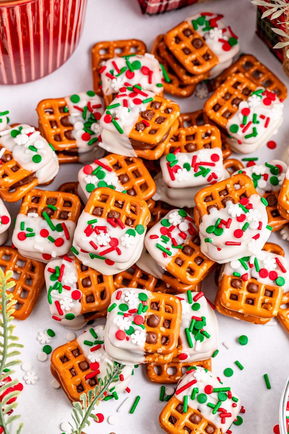 A pile of square pretzels crowned with white chocolate and festive red, green, and white sprinkles sits on a pristine surface.