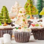 Christmas tree cupcakes are chocolate delights topped with frosting crafted to resemble festive evergreens. Each tree is adorned with colorful sprinkles and star-shaped toppings, while marshmallows are scattered nearby to complete the holiday scene.