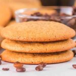 A stack of three brown coffee cookies surrounded by scattered coffee beans, with a bowl of beans in the background.