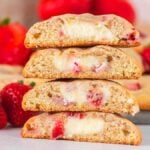 Three strawberry cheesecake cookies are stacked, with vibrant strawberries visible in the background.