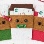 Three DIY felt Christmas gift card holders shaped like coffee cups in green, white, and red feature smiling faces and festive bows. They rest on a light background adorned with red beads and greenery, making them the perfect holiday touch.