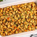 A baking dish brimming with golden-brown garlic and herb stuffing cubes, elegantly garnished with fresh herbs. A wooden spoon and a striped cloth napkin rest beside it, completing the cozy setup.