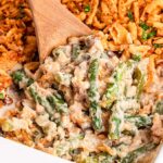 A wooden spoon expertly scoops a savory serving of green bean casserole, richly topped with crispy fried onions, from a pristine white baking dish.