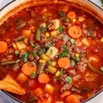 A hearty pot of hamburger soup loaded with carrots, potatoes, corn, and green beans. Topped with a sprinkle of parsley, a wooden spoon rests inside the pot.