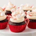 Hot chocolate cupcakes with white frosting, mini marshmallows, and small candy canes in red wrappers elegantly sit on a white stand.