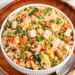 A bowl of leftover turkey fried rice with peas, carrots, corn, and green onions sits on a wooden plate, accompanied by a spoon.