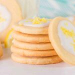 A stack of lemon shortbread cookies with white icing and lemon zest on top rests elegantly on a light surface, accented by a partially visible blue-patterned cloth in the background.