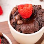 A chocolate lava cake mix in a white cup, topped with a raspberry and chocolate chips, creates the perfect mug cake delight. Additional raspberries are visible in the background, adding a vibrant touch to this indulgent treat.