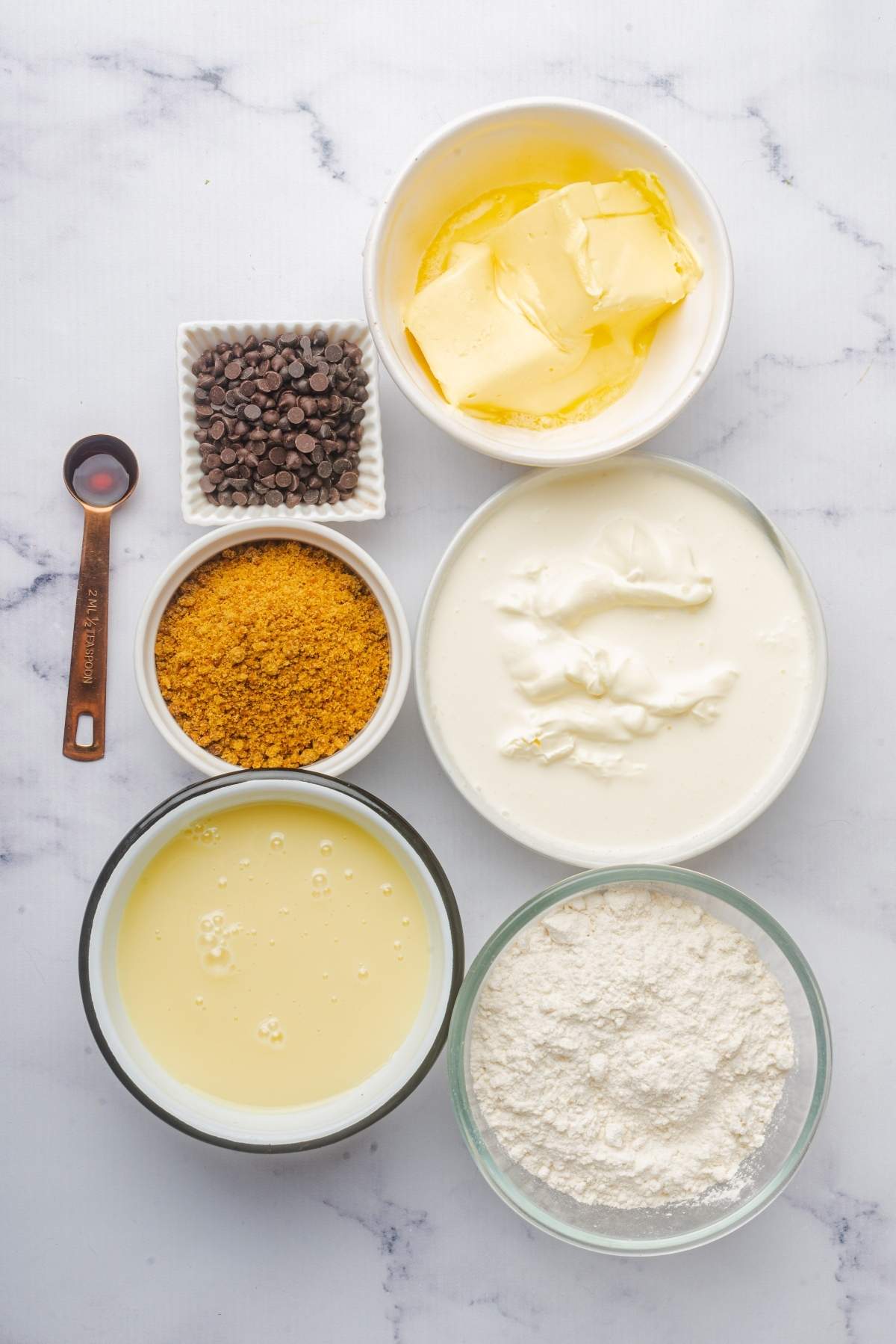 A top-down view reveals the essentials for no churn cookie dough ice cream: chocolate chips, butter, crushed graham crackers, sour cream, sweetened condensed milk, and flour—artfully arranged on a white marble surface.