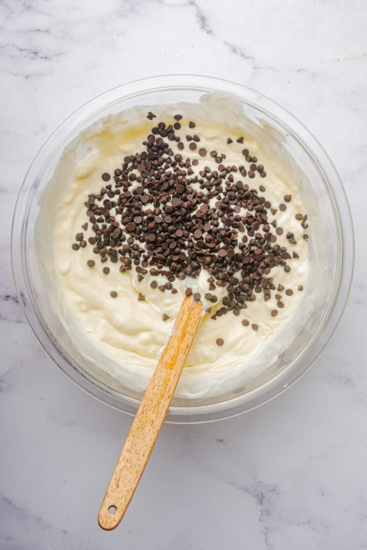 A bowl of creamy no churn cookie dough ice cream mixture with chocolate chips sprinkled on top, being gently stirred with a wooden spatula.