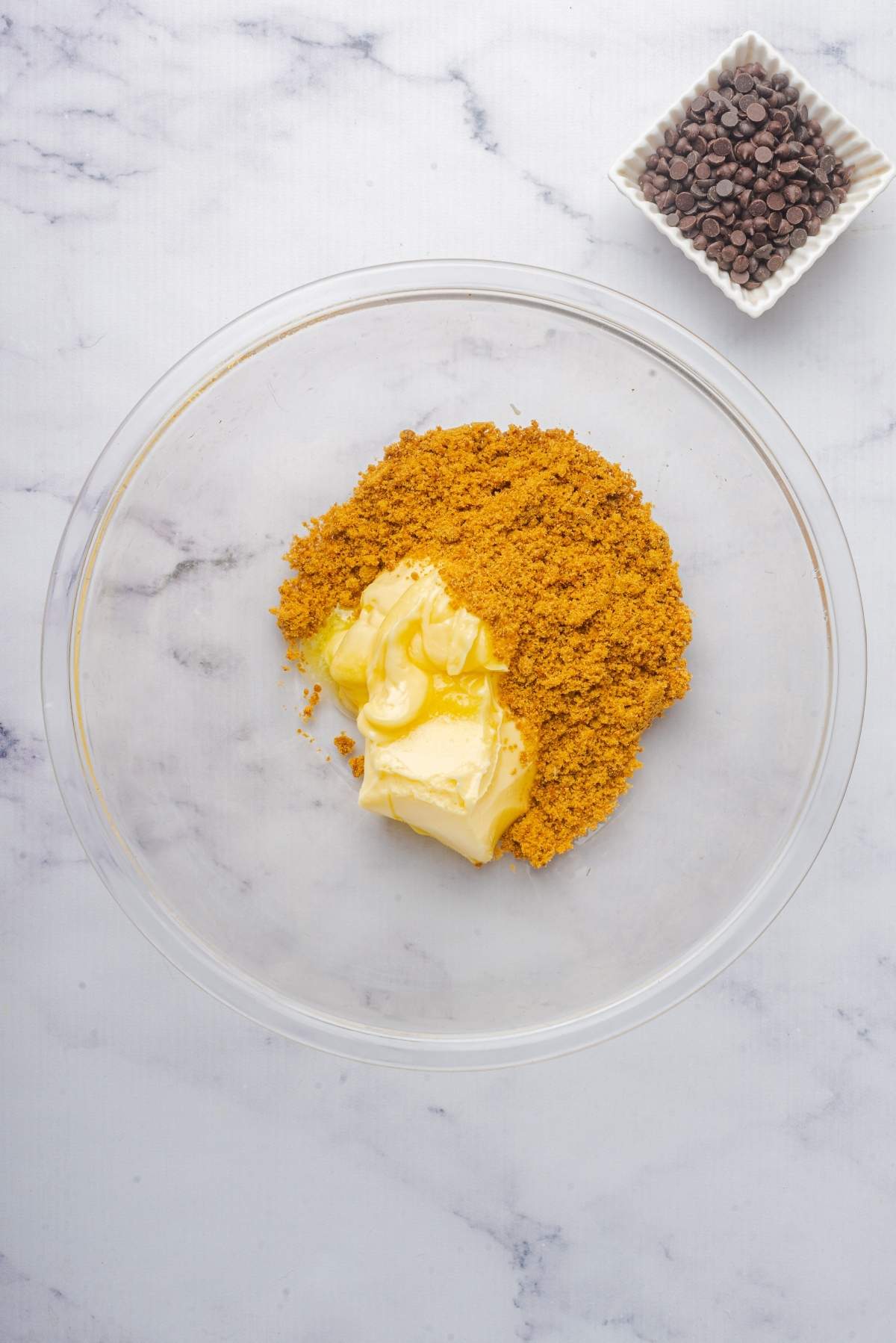 A glass bowl with butter, brown sugar, and honey sits ready to create a no churn cookie dough ice cream. Beside it, a small square dish filled with chocolate chips rests on the marble surface.