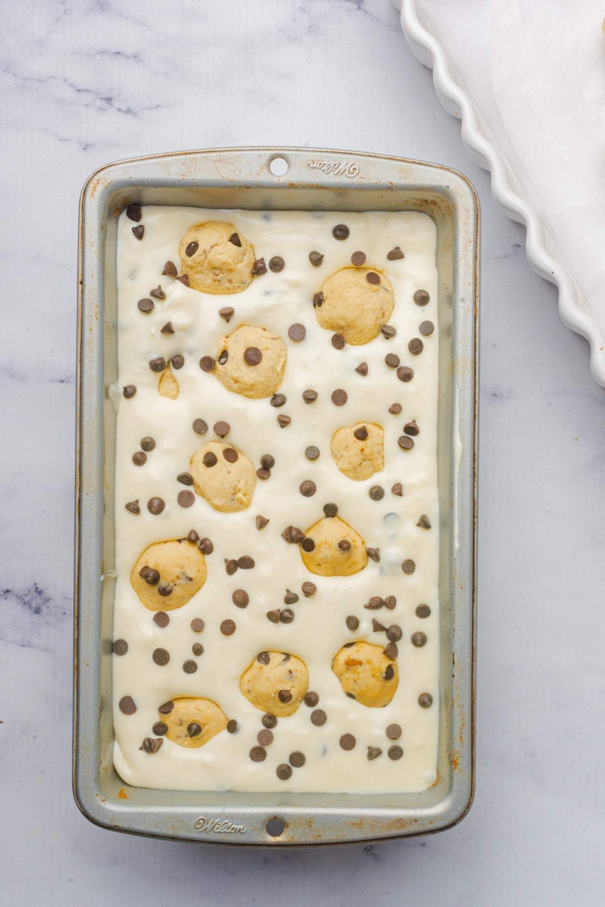 A baking pan with cheesecake batter topped with chocolate chip cookie dough balls and scattered chocolate chips, bringing to mind the delicious fusion found in no churn cookie dough ice cream.