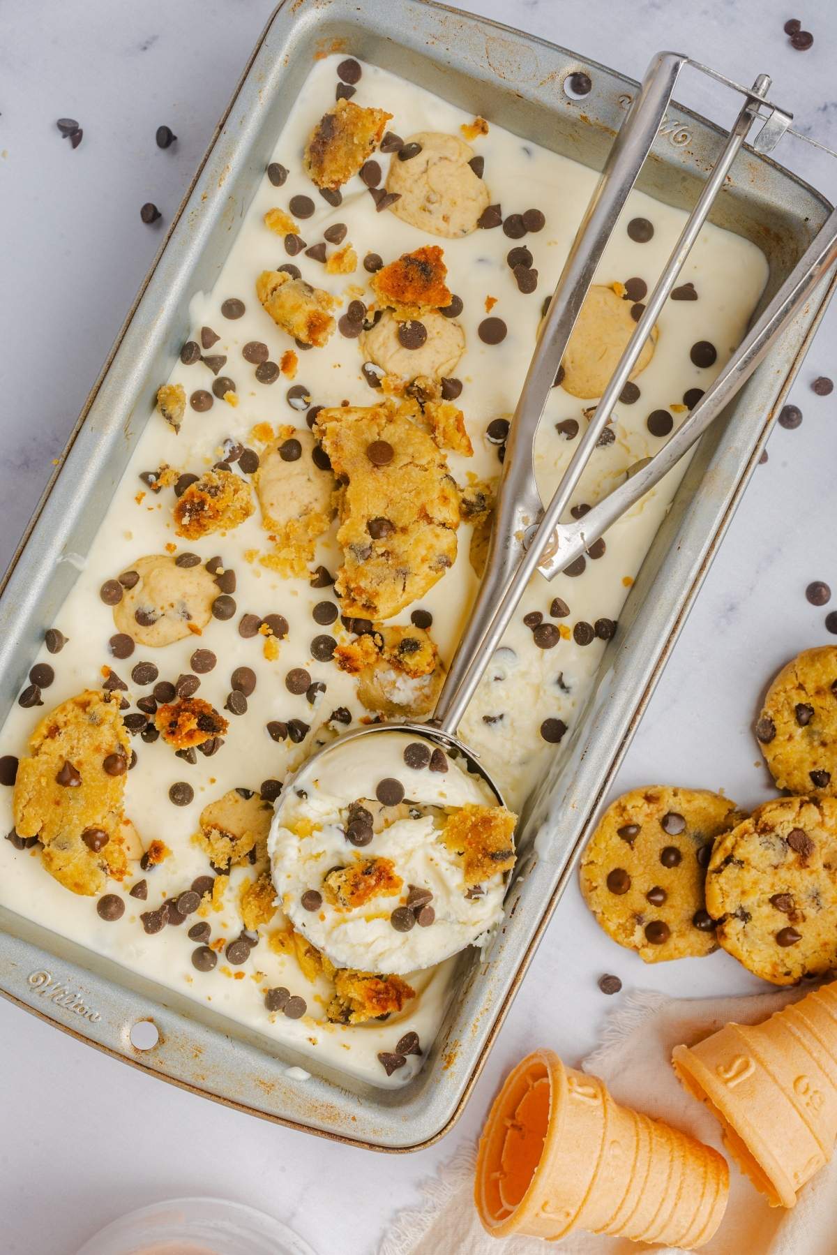 A tray of no churn cookie dough ice cream with chocolate chips and cookie chunks. A scoop is partially submerged in the creamy delight. Discarded ice cream cones and cookies are nearby, hinting at a delicious indulgence.