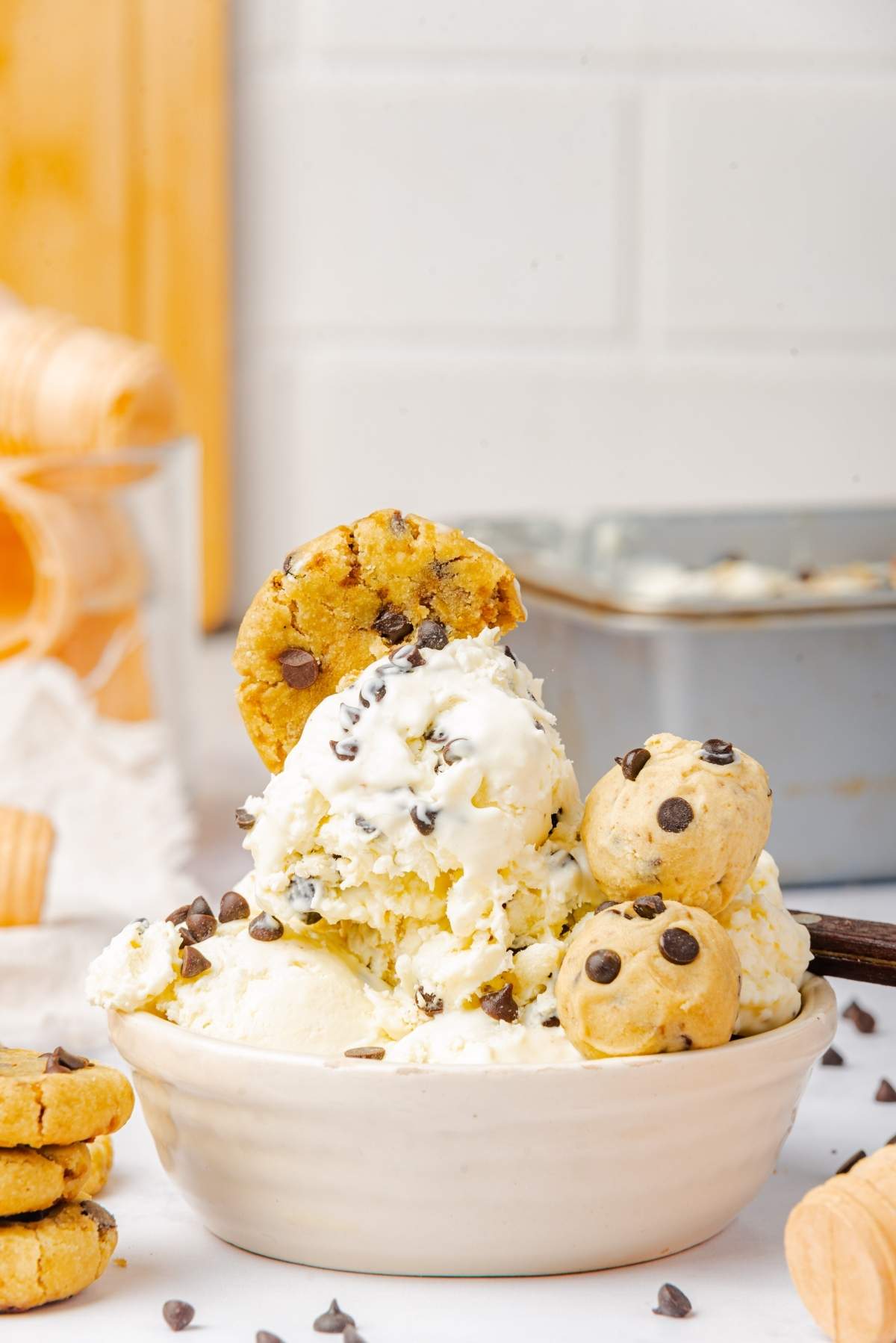 A bowl of no churn cookie dough ice cream with chocolate chips, topped with a cookie, surrounded by cookie balls and extra chocolate chips on a table.