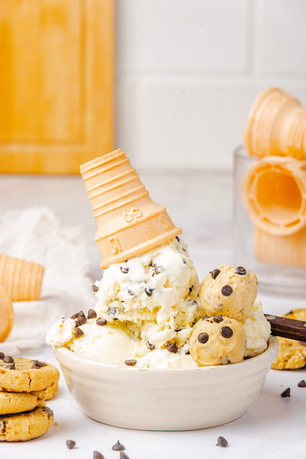 A bowl of no churn cookie dough ice cream studded with chocolate chips and cookie dough balls, crowned with an upside-down ice cream cone. Cookie pieces and more cones are scattered in the background, adding to the sweet temptation.