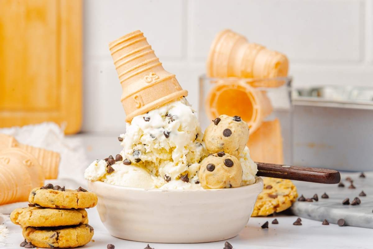 A bowl of no churn cookie dough ice cream topped with a waffle cone sits amidst scattered chocolate chip cookies and empty cones.