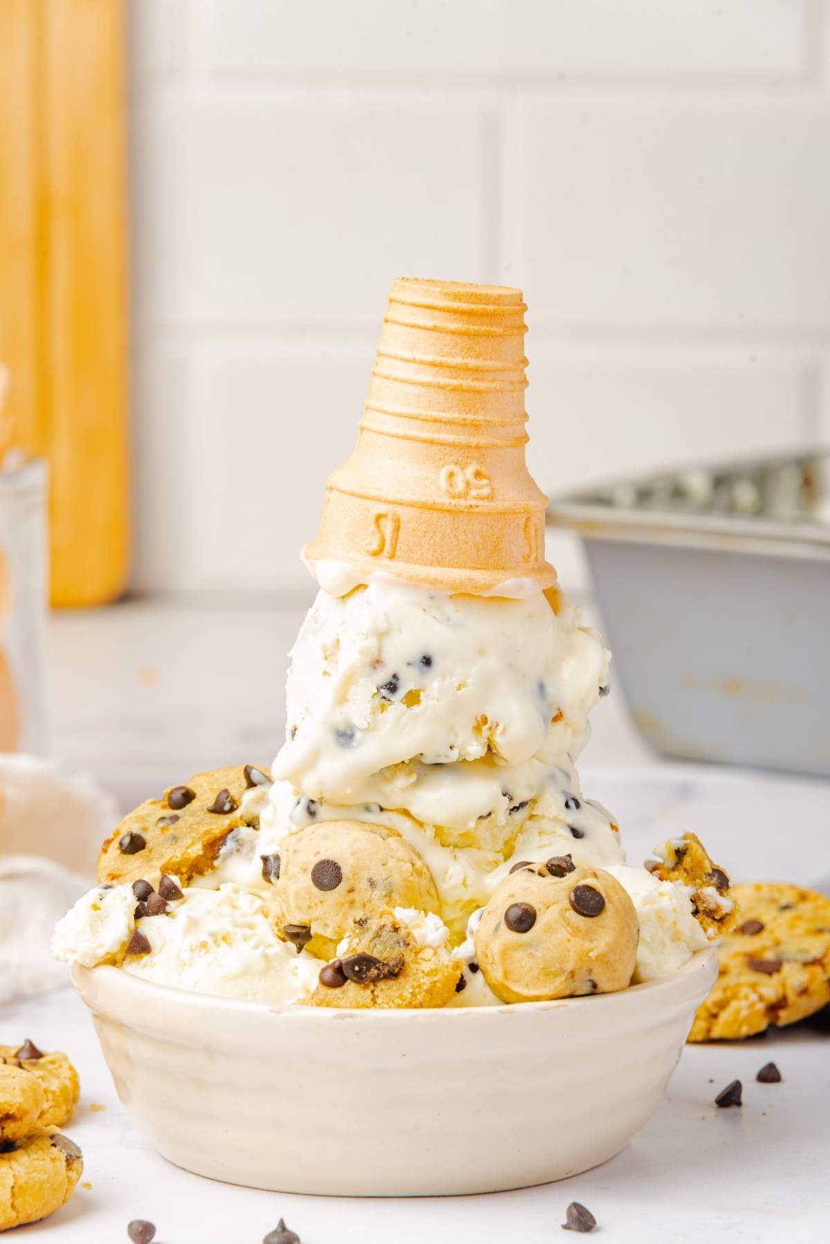 No churn cookie dough ice cream topped with chocolate chip cookies and a wafer cone, served in a bowl on a white surface.