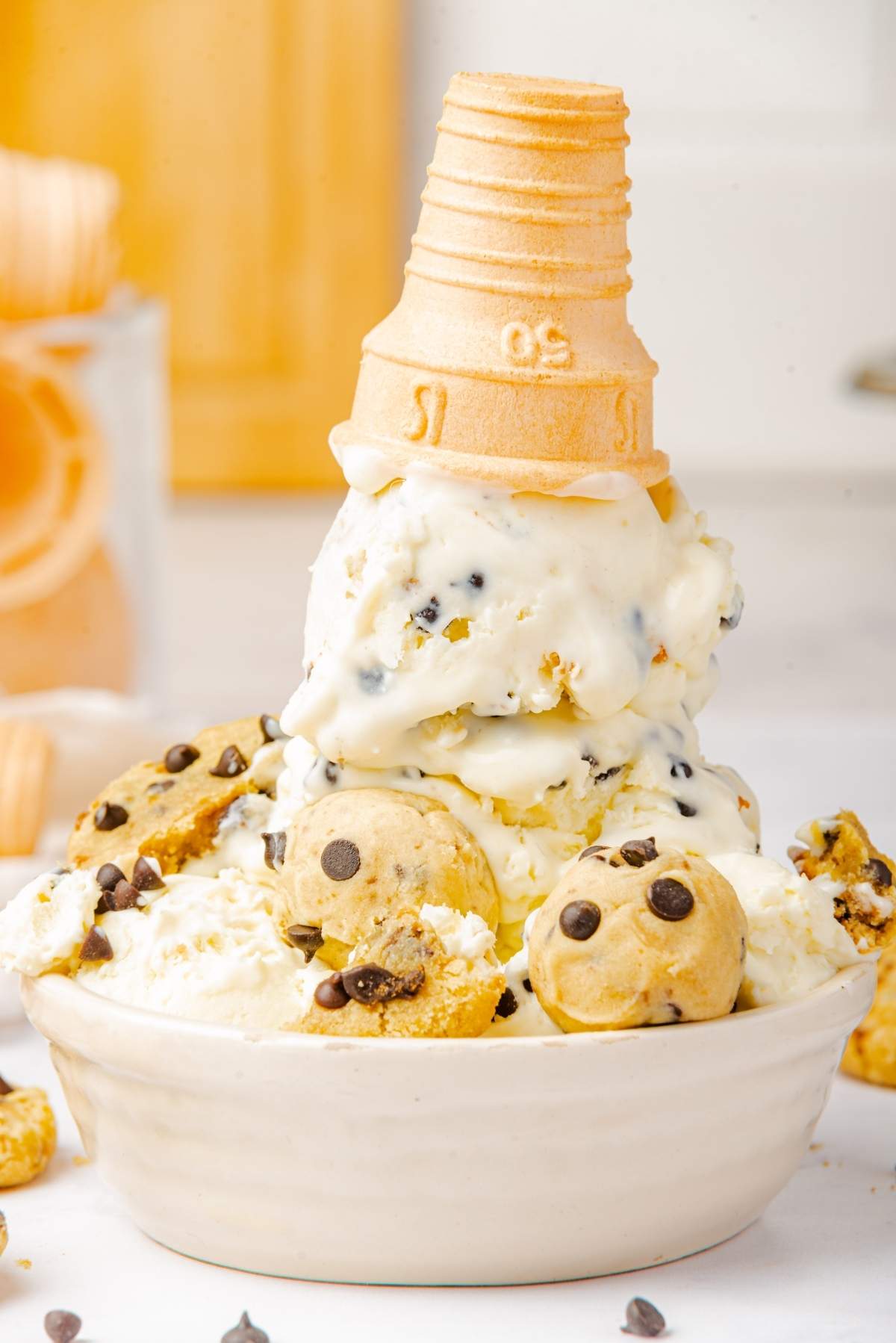 A bowl of no churn cookie dough ice cream is topped with an upside-down ice cream cone. Cookie dough pieces and chocolate chips are scattered around the bowl.