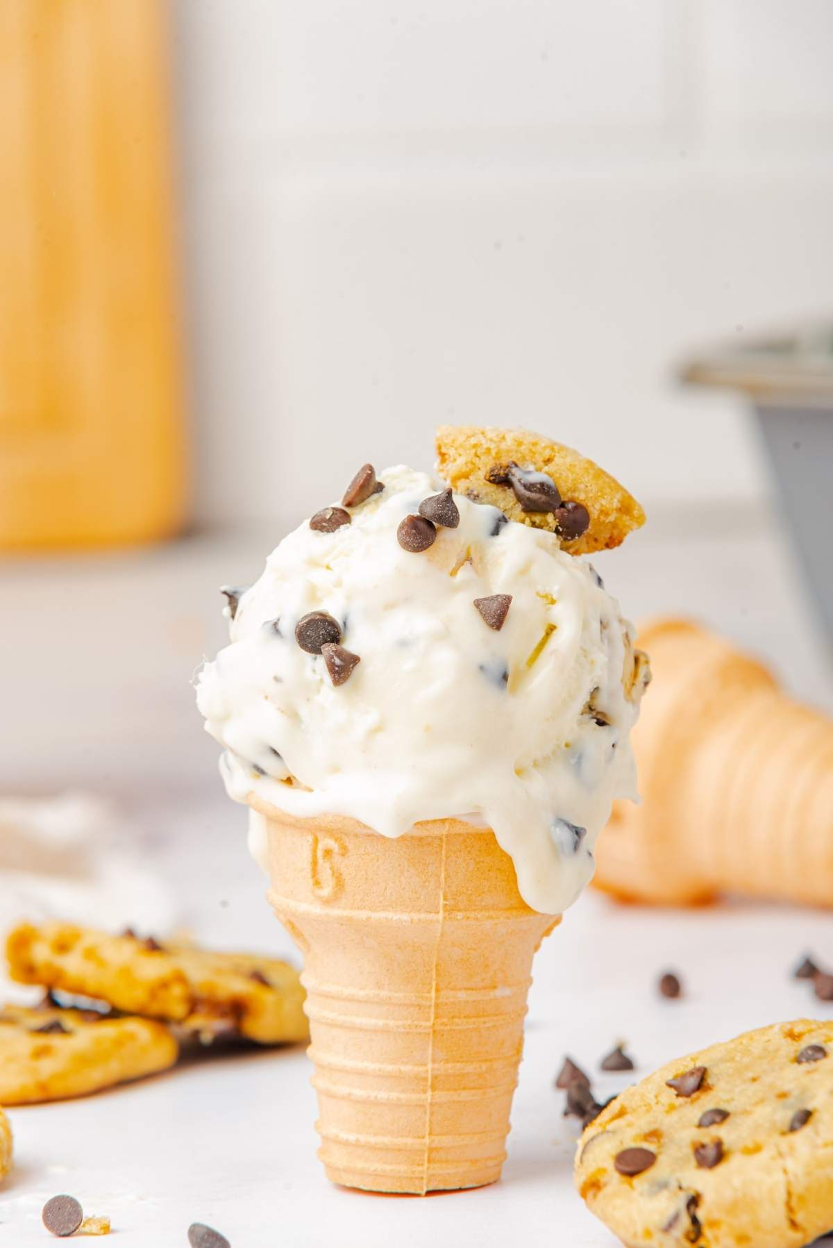 A delightful no churn cookie dough ice cream cone features a scoop of creamy goodness, crowned with chocolate chips and a mini cookie, all nestled amidst a scattering of broken cookies.