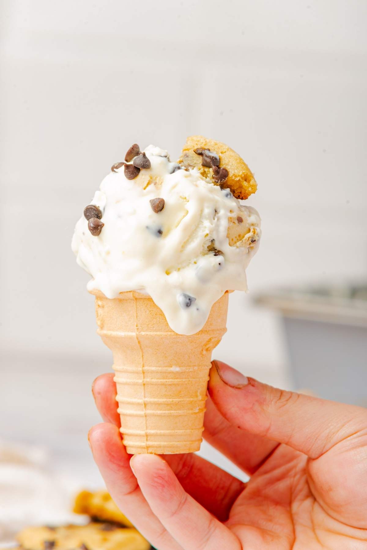 A hand holds a cone of vanilla ice cream speckled with chocolate chips and crowned with a cookie piece, reminiscent of no-churn cookie dough ice cream.