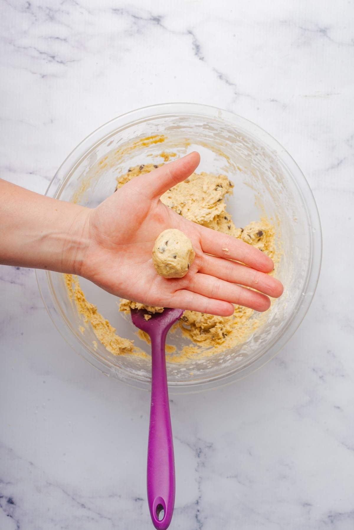 A hand holds a ball of unbaked cookie dough, ready to become no churn cookie dough ice cream, over a clear bowl and purple spoon on a marble surface.