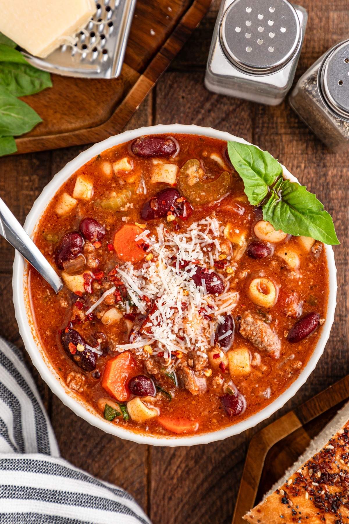A bowl of hearty minestrone, reminiscent of pasta e fagioli soup, brimming with beans, vegetables, and grated cheese, garnished with basil. A spoon rests in the bowl while salt, pepper shakers, and bread sit enticingly nearby.
