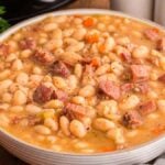 A bowl of hearty slow cooker ham and bean soup brimming with chunks of tender meat, white beans, and vibrant vegetables. An onion and fresh green herbs rest invitingly beside the bowl.