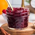 A small glass jar of spiced cranberry orange jam with a gold spoon, surrounded by cinnamon sticks, orange slices, and a green cloth.